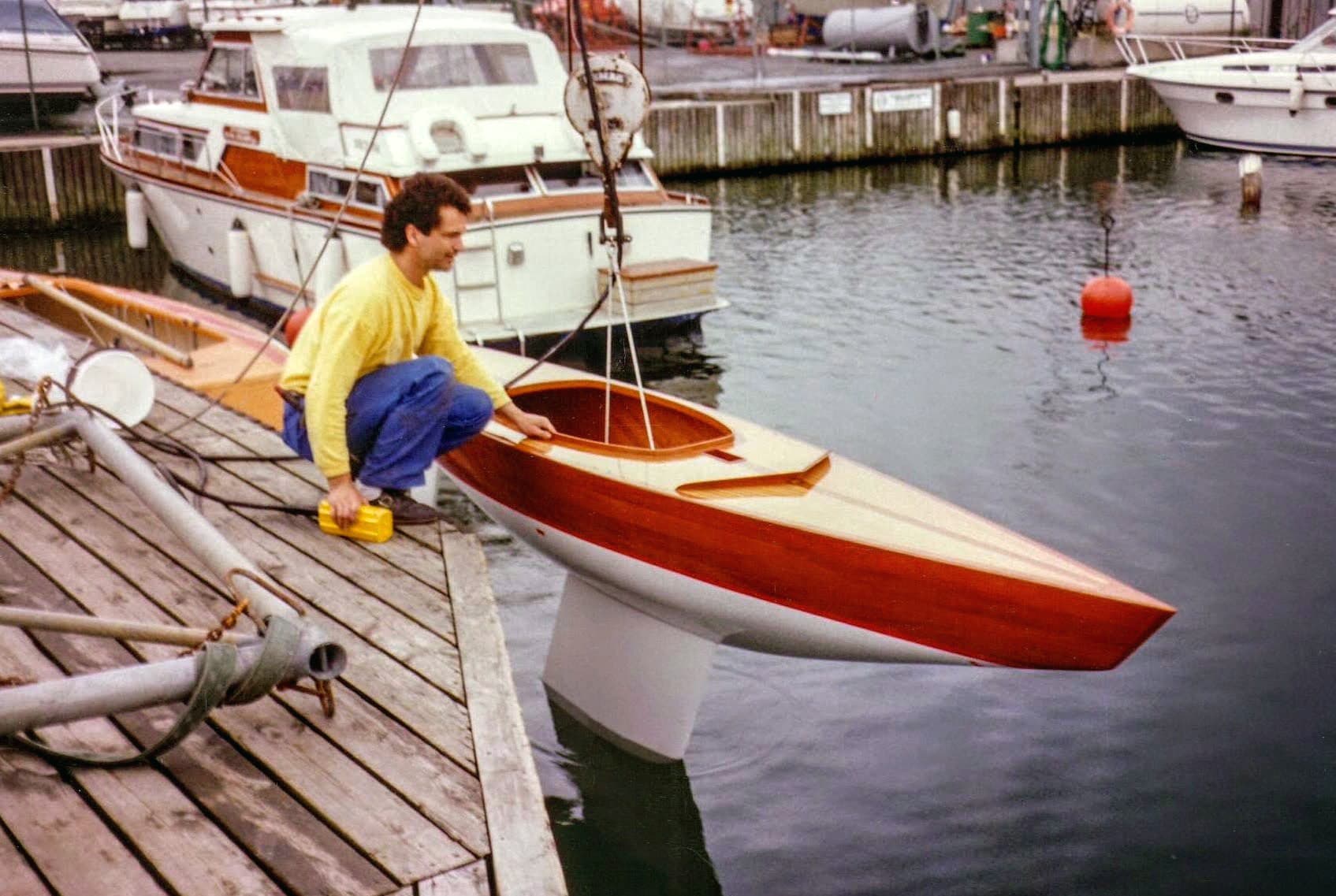 A varnished wooden Norlin 2.4mR alongside a dock, steadied by hand as it is lowered into the water.